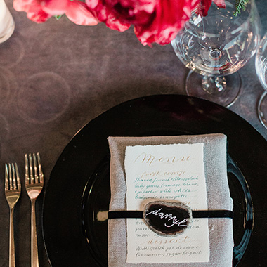 close up of table setting: copperplate calligraphy menu in gold and green wrapped with a black ribbon. On it is a flat agate with darryl written on it in white